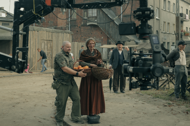 Rudowłosa starsza kobieta w brązowej sukni z koszem wiklinowym w ręku, obok mężczyzna z koszem pomarańczy otoczeni urządzeniami filmowymi