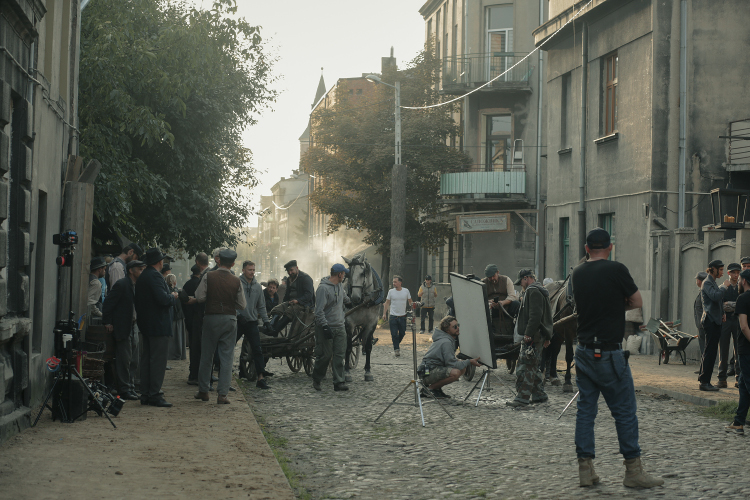 Wybrukowana kamieniami ulica pośród kamienic, aktorzy i filmowcy przygotowujący się do zdjęć, na środku wóz z koniem