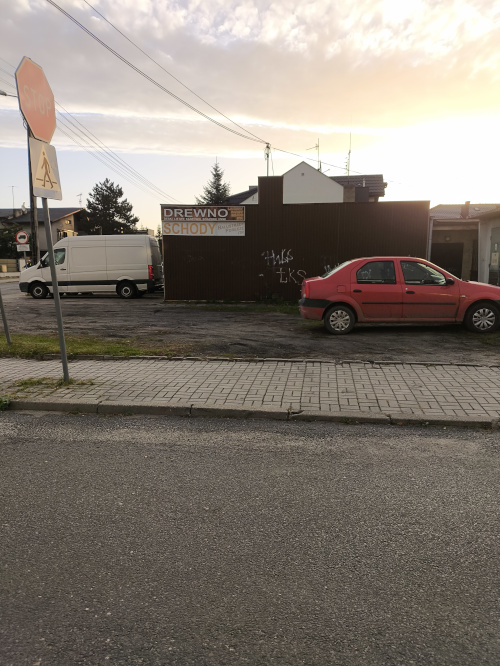 Czerwony samoch&oacute;d i biała furgonetka przed płotem z reklamą firmy stolarskiej. Obok chodnik, ulica i znak STOP.