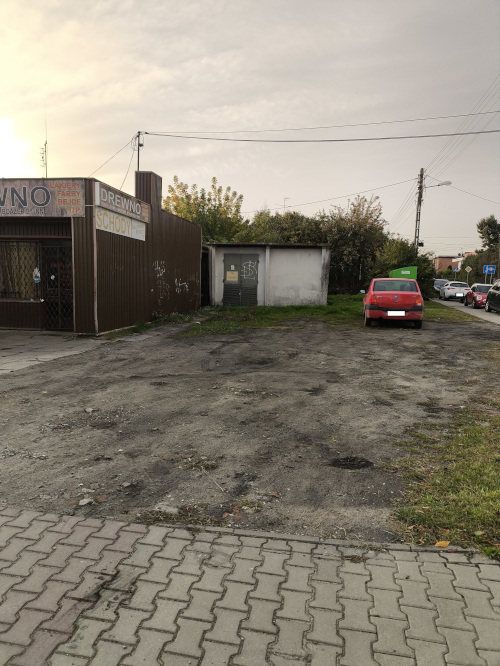 Gruntowy plac parkingowy obok brązowego budynku z szyldami. Po prawej czerwony samoch&oacute;d. Na pierwszym planie chodnik.