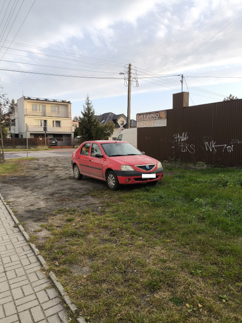 Czerwony samoch&oacute;d zaparkowany na gruntowym placu przy płocie z reklamą. W tle dom jednorodzinny i słup elektryczny.