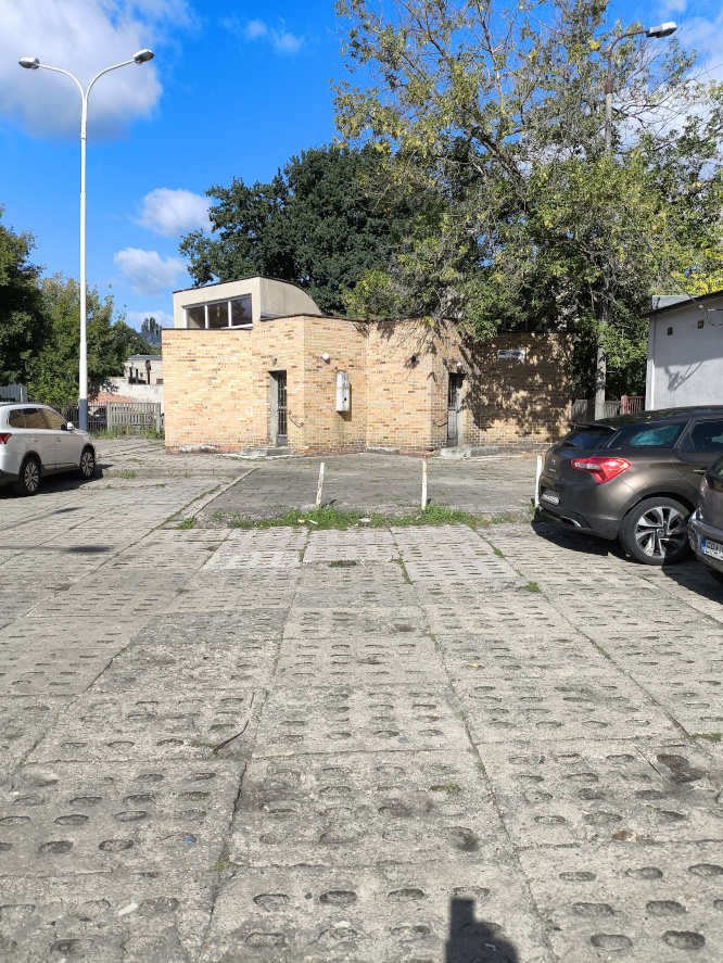 Betonowy plac parkingowy z samochodami przed niskim budynkiem z jasnej cegły. W tle drzewa i słoneczne niebo
