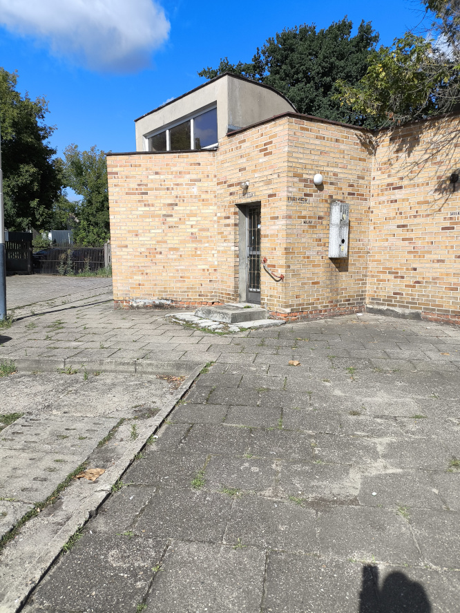 Zbliżenie na niski budynek z jasnej cegły z przeszkloną nadbud&oacute;wką i drzwiami. Z przodu chodnik z betonowych płyt.