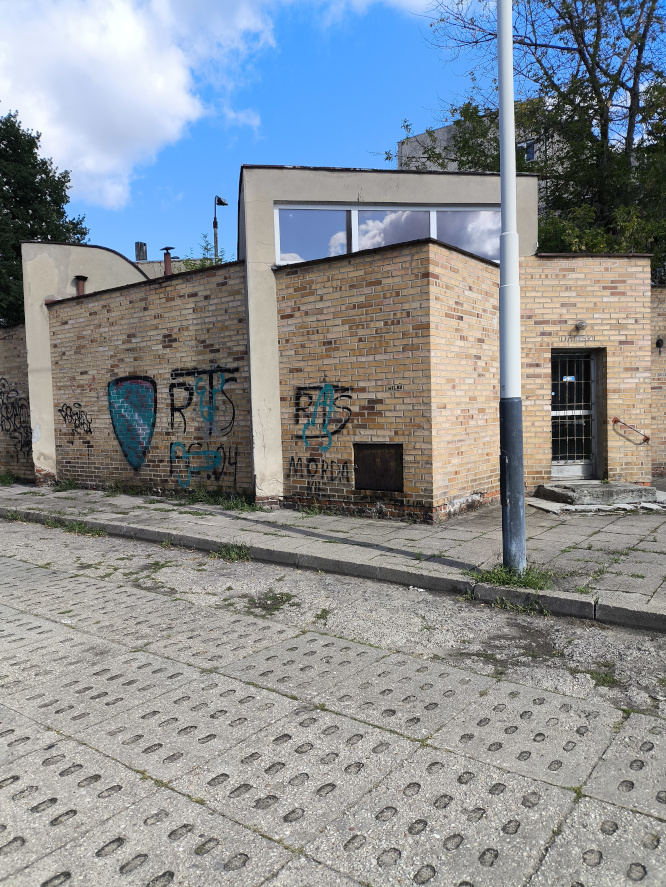 Ceglany, niski budynek z graffiti na ścianach i słupem oświetleniowym na pierwszym planie. Przed nim betonowy plac.