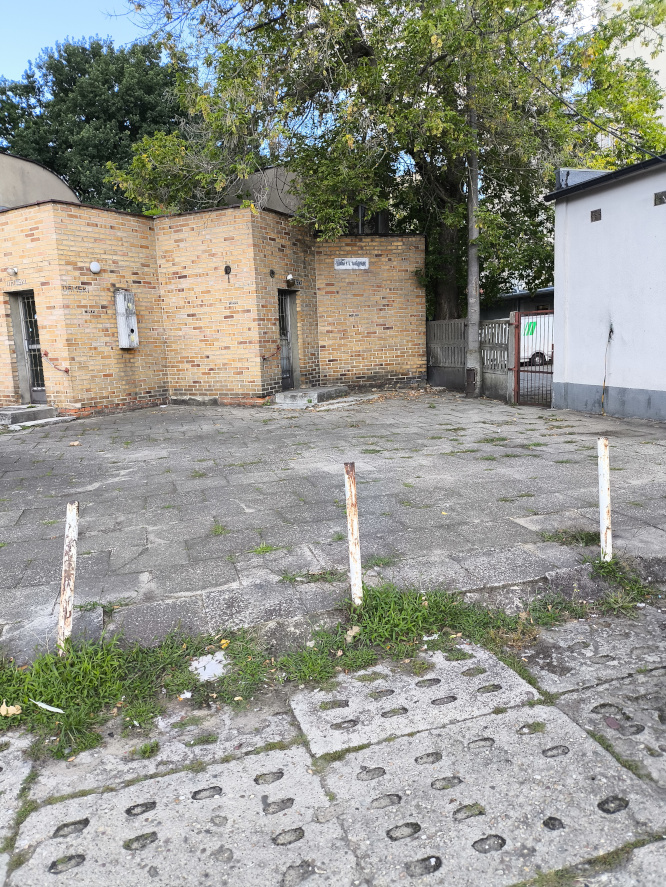 Betonowy plac z metalowymi słupkami na pierwszym planie, prowadzący do niskiego budynku z jasnej cegły otoczonego drzewami.