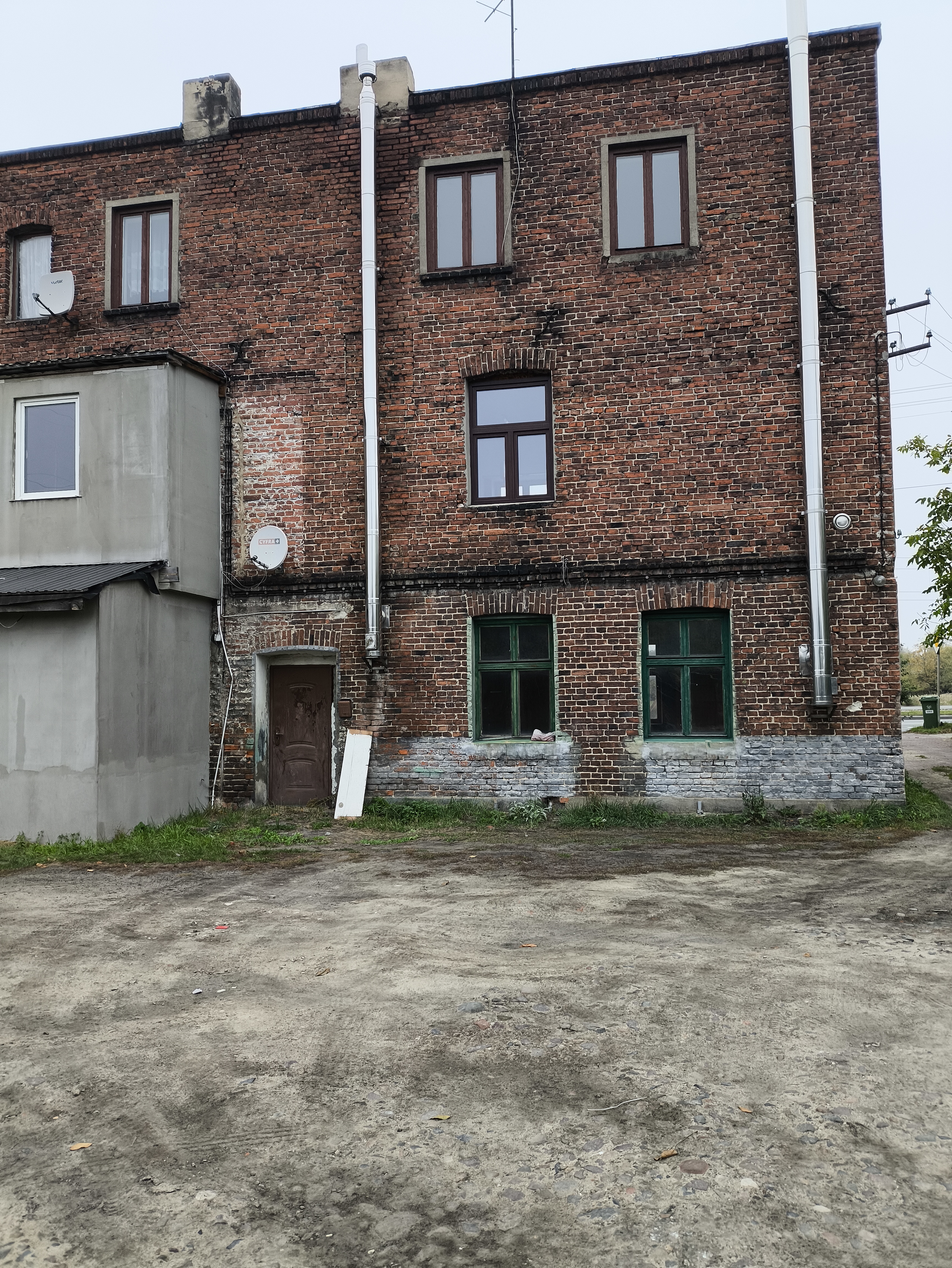 Ceglany, wielokondygnacyjny budynek z kilkoma oknami i rurami spustowymi, widok od strony podw&oacute;rza z nieutwardzonym placem.