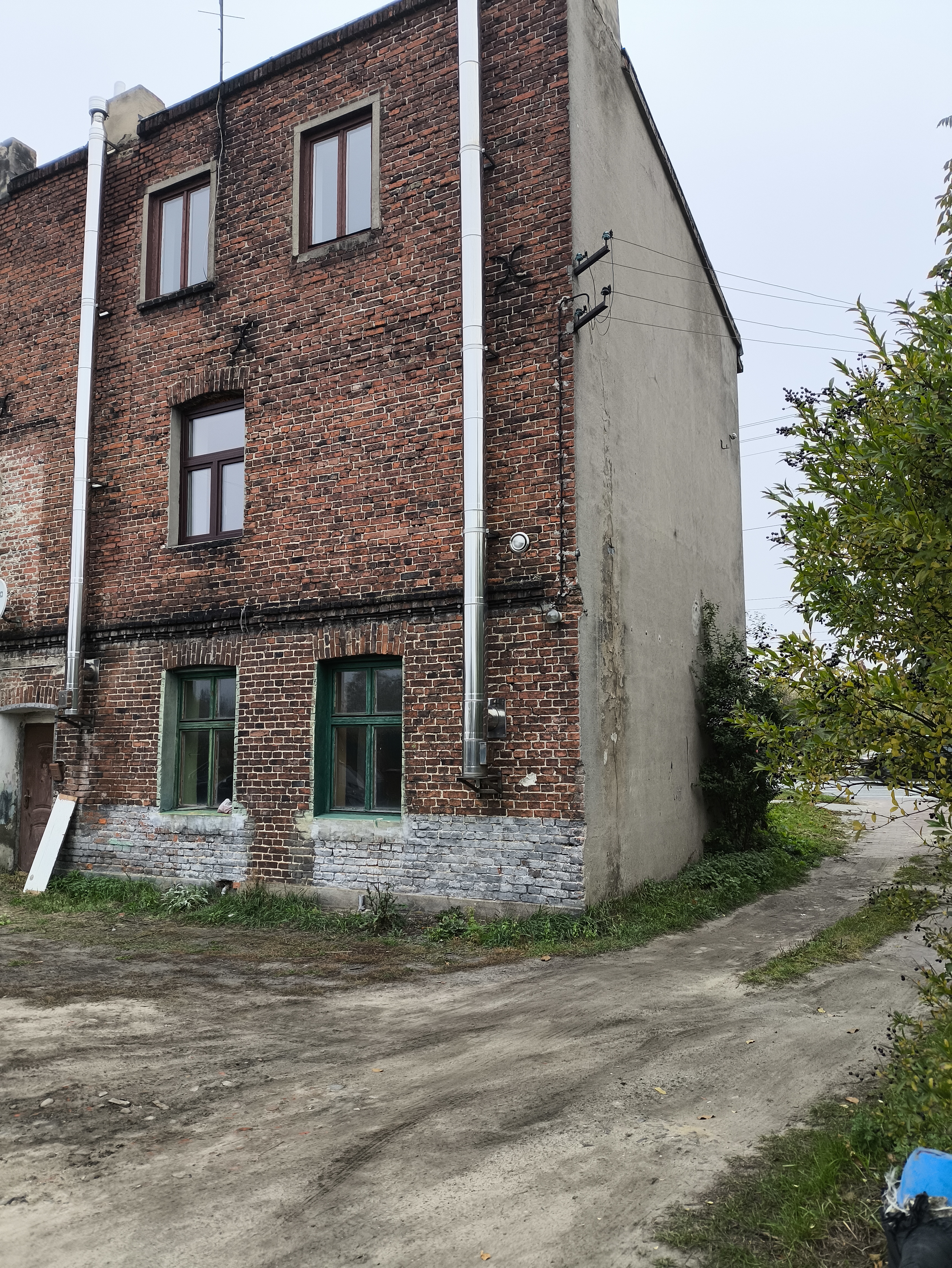  Ceglany budynek wielokondygnacyjny od strony bocznej, z oknami i rurami spustowymi, przy nieutwardzonej drodze i roślinności.