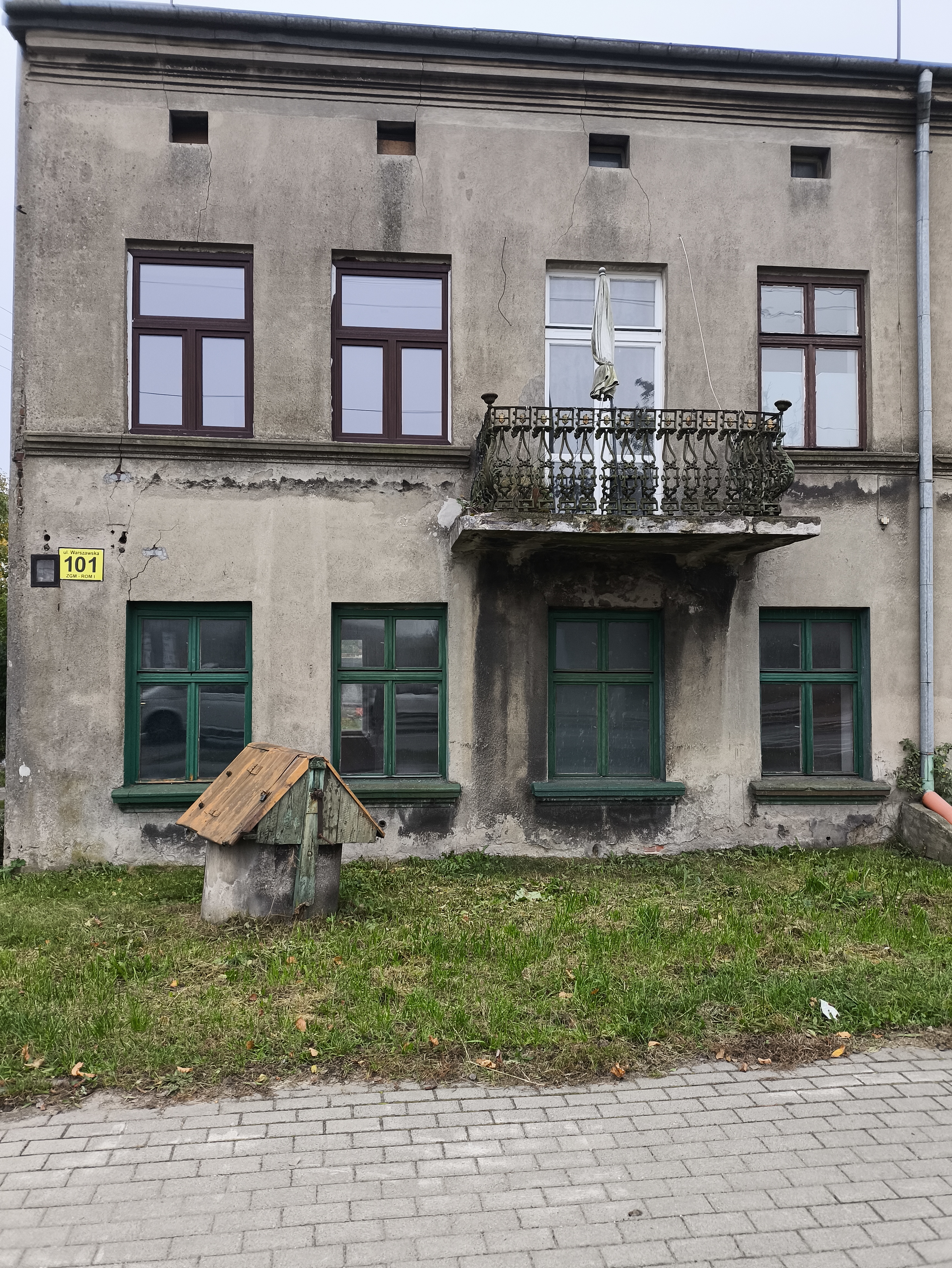 Dwukondygnacyjny budynek z tynkowaną elewacją, balkonem nad wejściem i zieloną stolarką okienną oraz drzwiową.