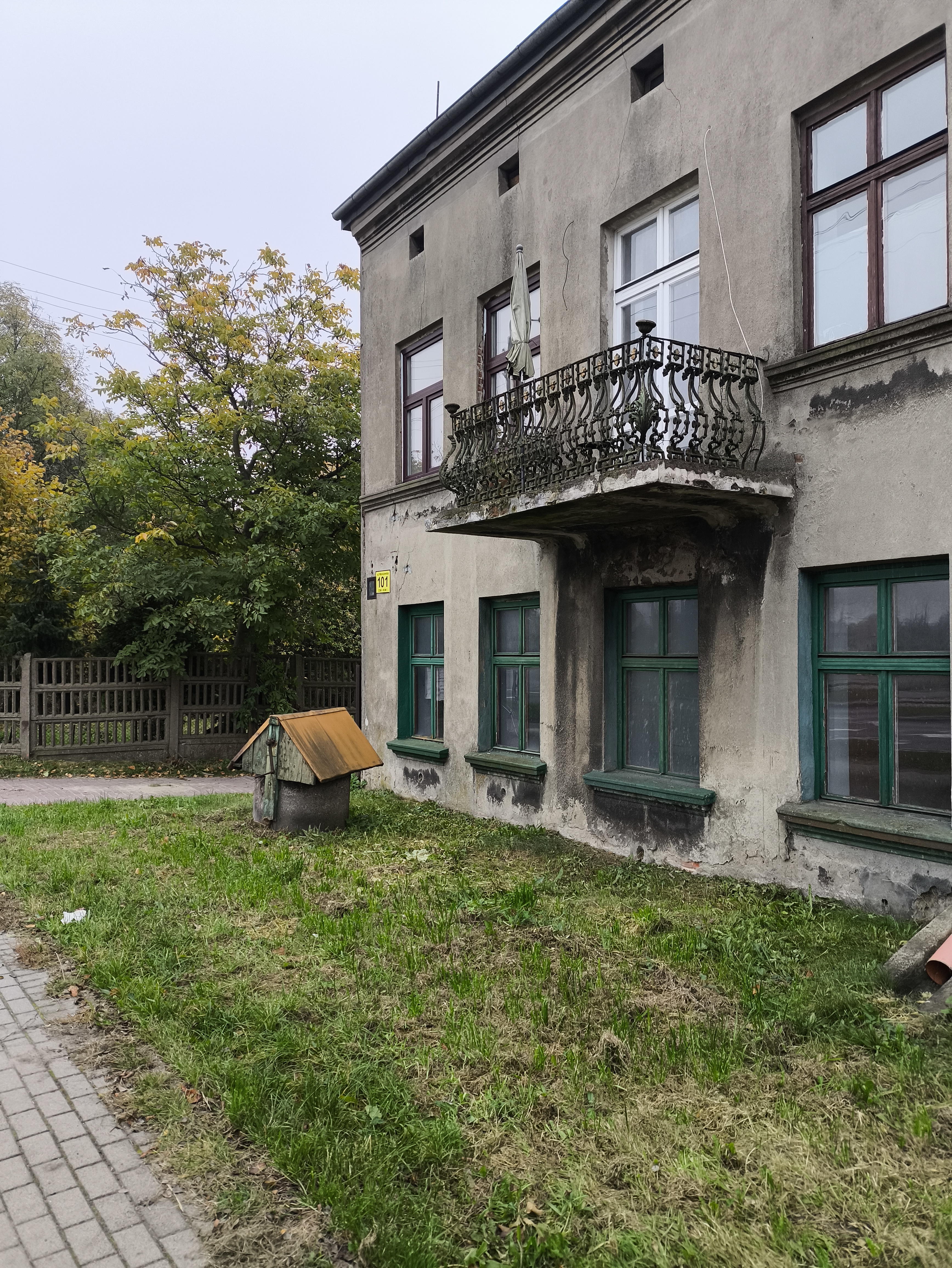  Fragment budynku z tynkowaną elewacją i balkonem nad wejściem, otoczony trawnikiem i drzewami