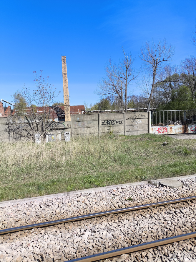Widok z torowiska na betonowy płot z graffiti, za kt&oacute;rym stoi wysoki komin i stare budynki fabryczne pod bezchmurnym niebem
