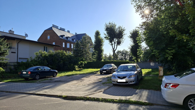 Plac gruntowy i brukowany przy budynkach mieszkalnych w słoneczny dzień. Kilka zaparkowanych samochod&oacute;w pod drzewami.