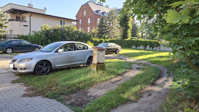 Srebrny samoch&oacute;d zaparkowany na trawniku obok skrzynki elektrycznej. W tle budynki mieszkalne oraz zieleń.