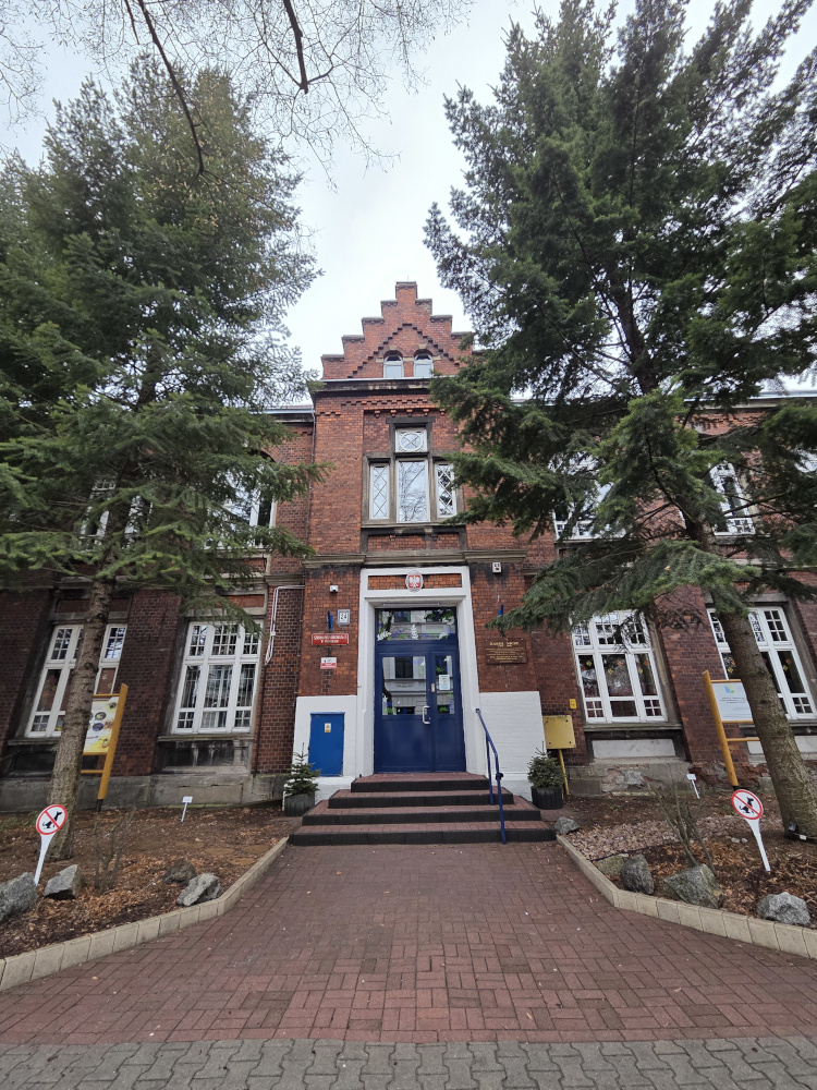 Wejście do ceglanego, neogotyckiego budynku z niebieskimi drzwiami i schodkowym szczytem. Po bokach rosną wysokie świerki, a przed nimi biegnie chodnik.