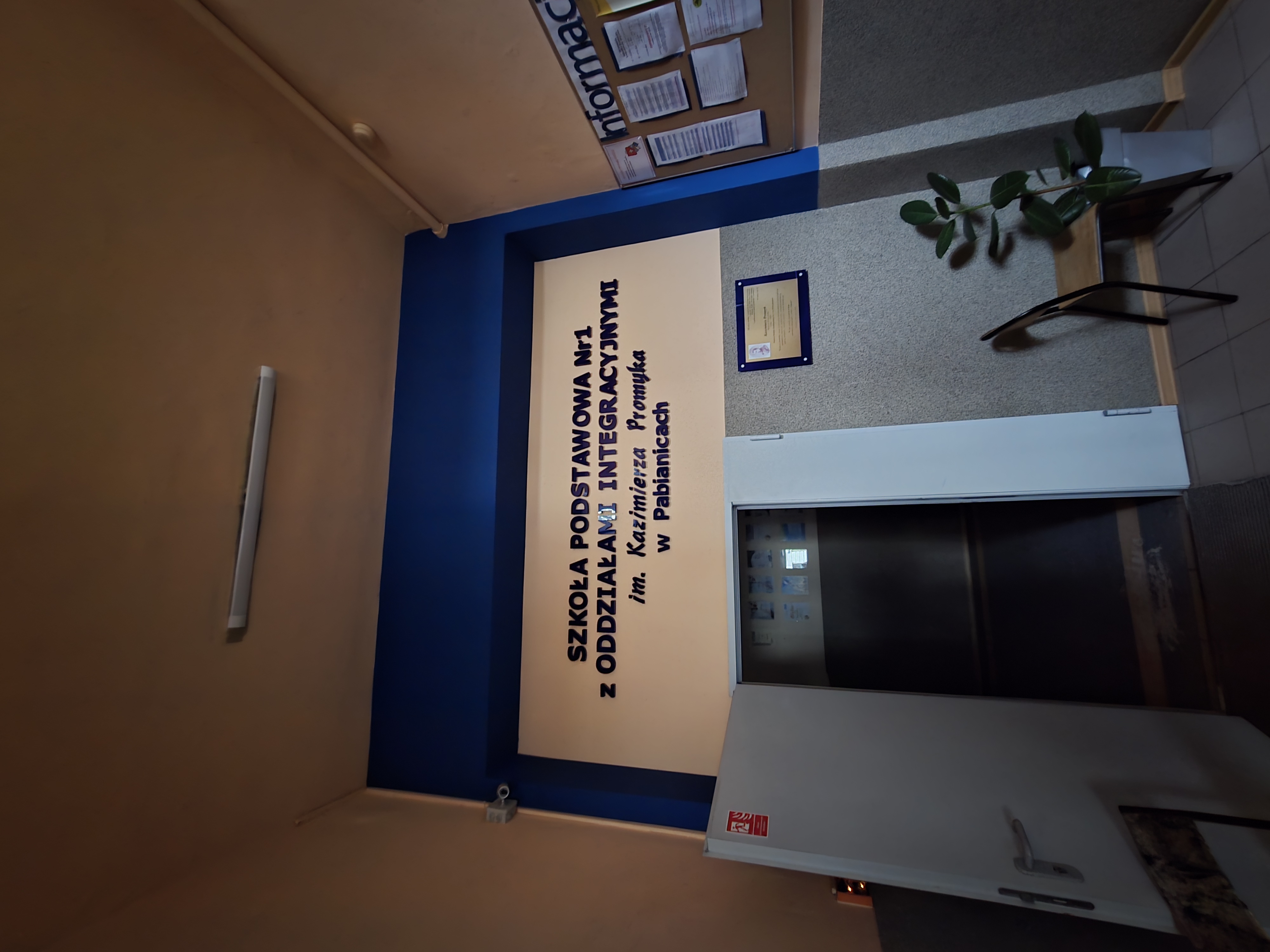 Wnętrze korytarza szkoły w Pabianicach. Nad drzwiami napis: Szkoła Podstawowa nr 1 im. Kazimierza Promyka. Obok tablica informacyjna i krzesło z kwiatem.