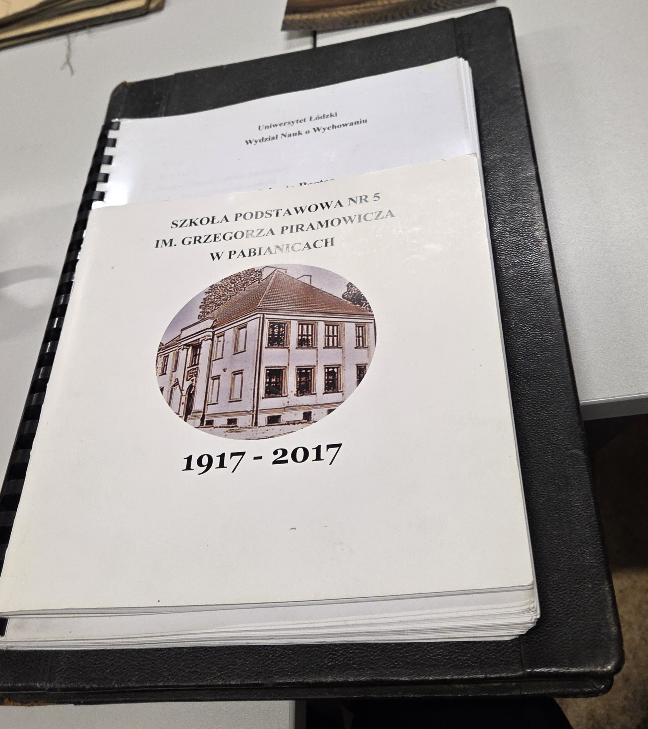 Okładka publikacji jubileuszowej Szkoły Podstawowej nr 5 w Pabianicach (1917&ndash;2017) ze zdjęciem budynku, leżąca na czarnej teczce.