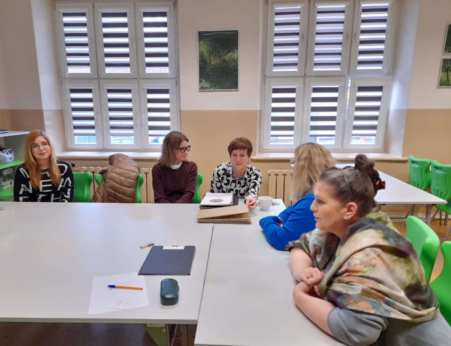 Grupa pięciu kobiet siedzi przy jasnym stole w sali konferencyjnej. Rozmawiają, trzymając przed sobą dokumenty. W tle widać okna z pasiastymi roletami.