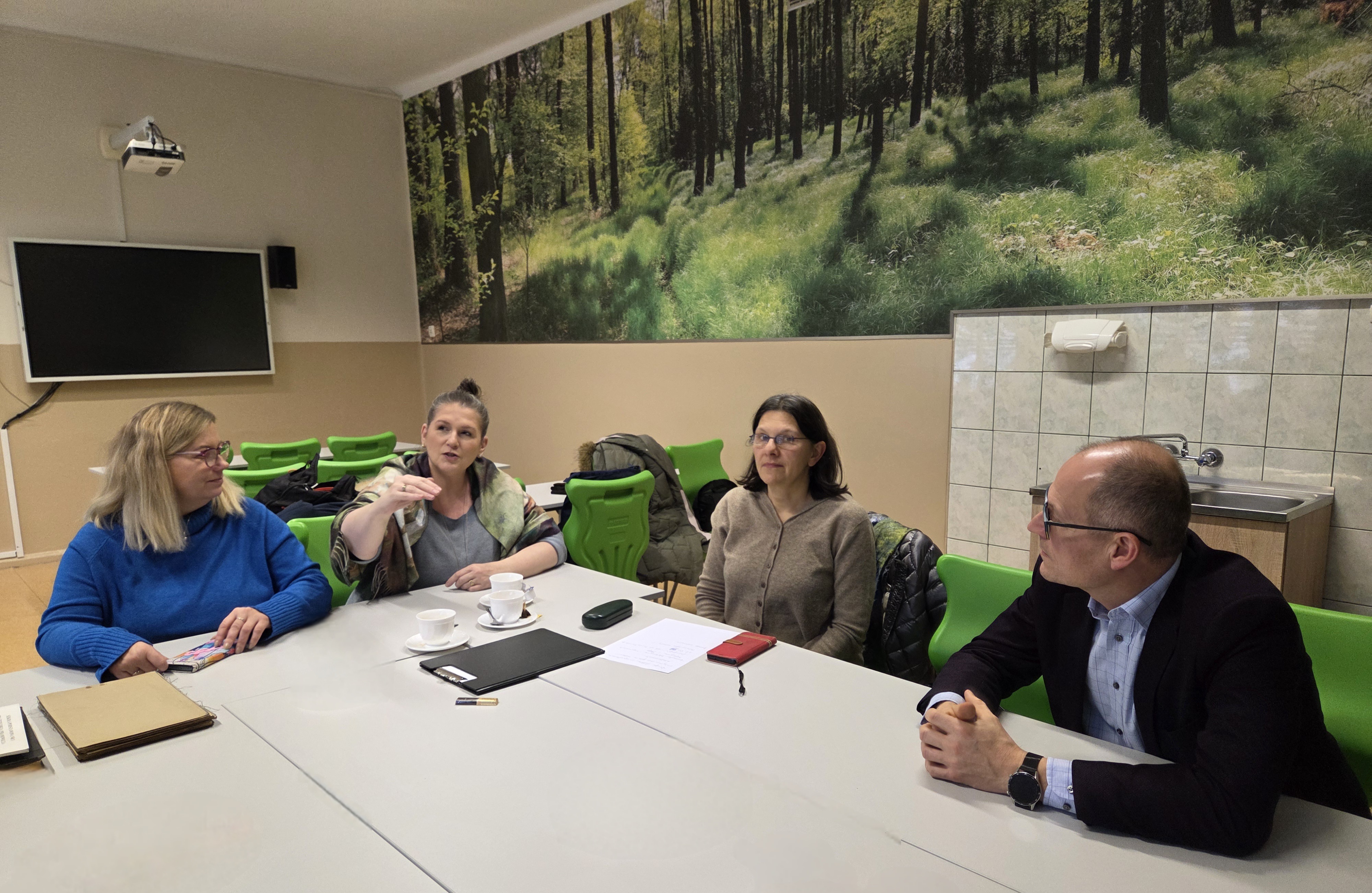 
Trzy kobiety i mężczyzna siedzą przy stole podczas spotkania. Na ścianie w tle fototapeta z lasem. Przed uczestnikami leżą notatki i filiżanki kawy.