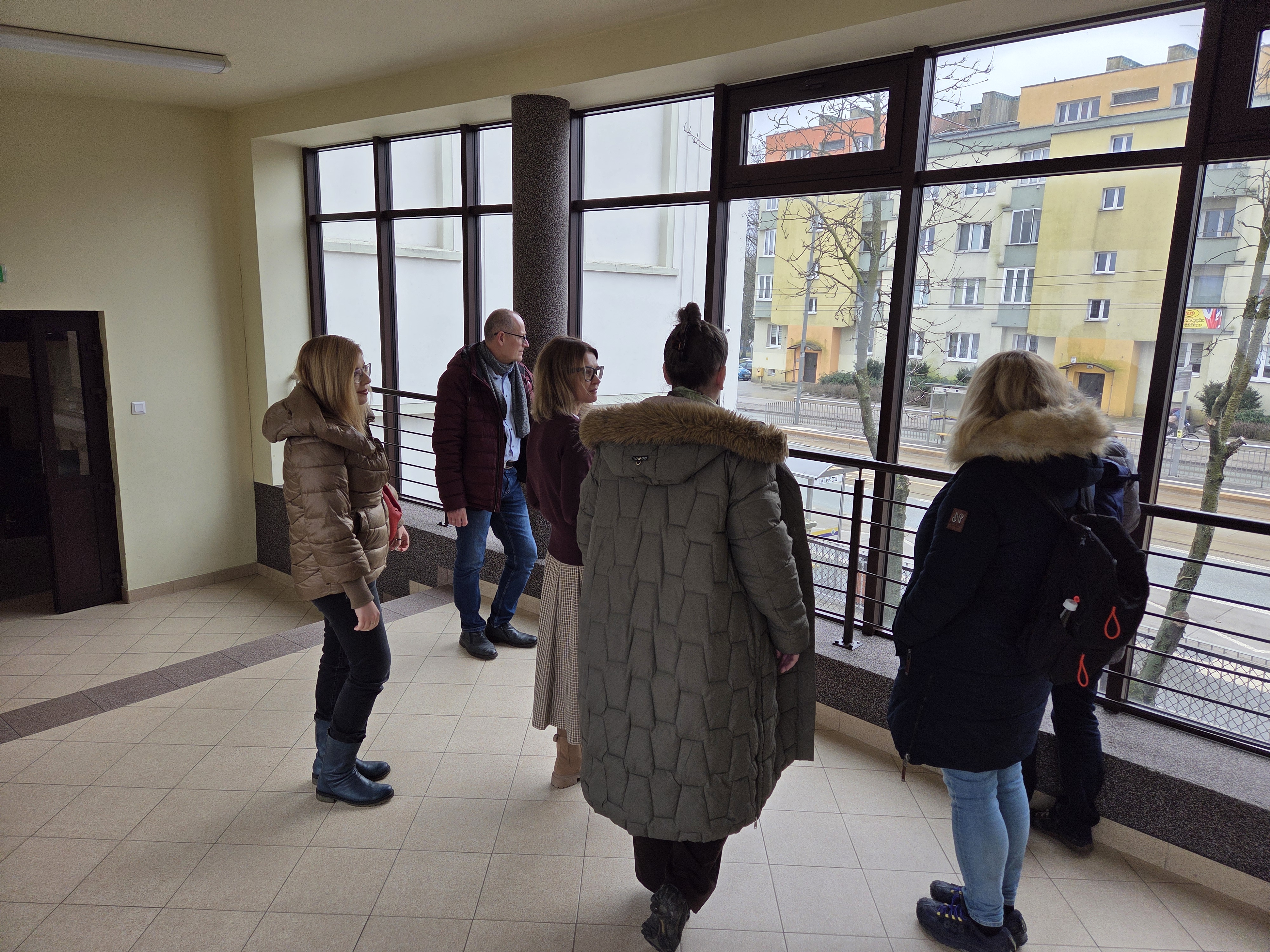 Grupa os&oacute;b w ubraniach wierzchnich stoi na przeszklonym korytarzu i spogląda przez duże okno na ulicę oraz bloki mieszkalne.